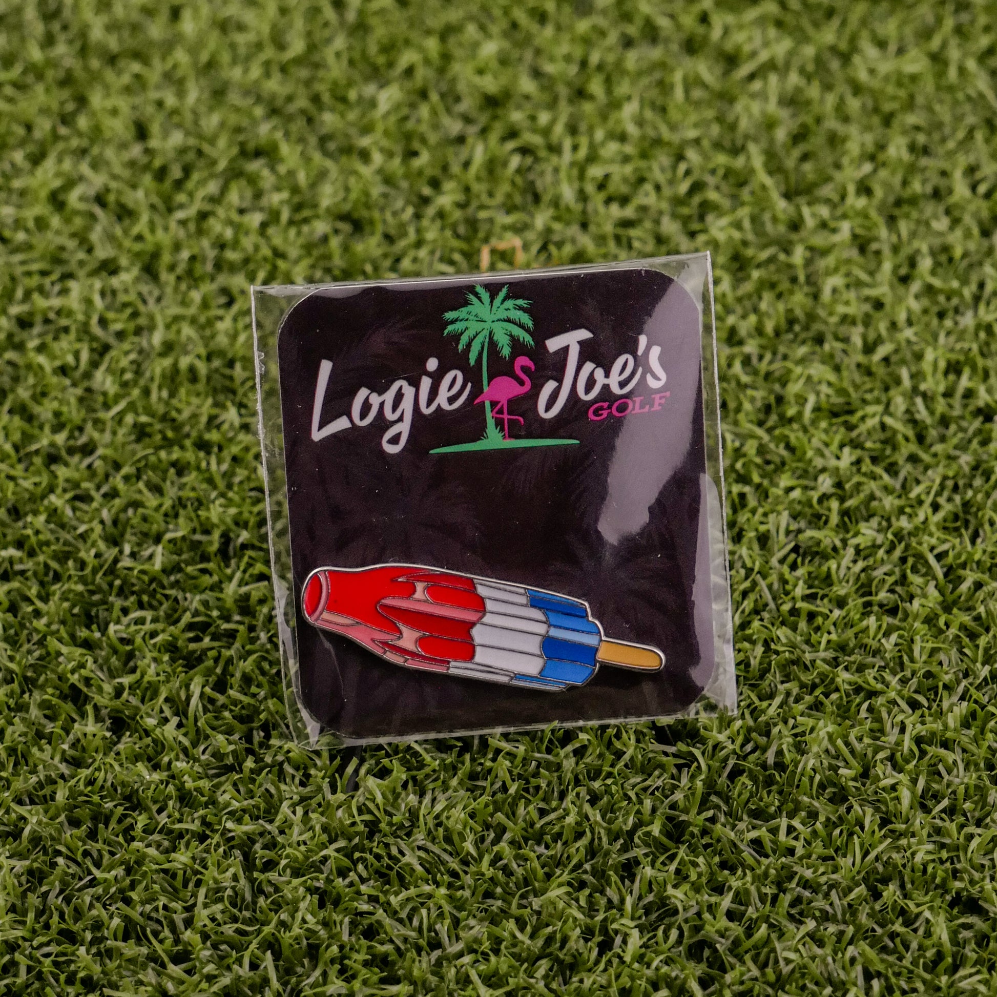 The Red White Blue Popsicle by Ballmarkers by Logie Joe’s is a novelty golf pin that doubles as a premium ball marker, shaped like a rocket pop and displayed on black packaging with artificial green grass.