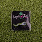 The Sperm Cell, a premium ball marker from Ballmarkers by Logie Joes, features cartoon eyes and comes on artificial grass with a black card in white, green, and pink plus flamingo and palm tree—funny golf accessories at their best.