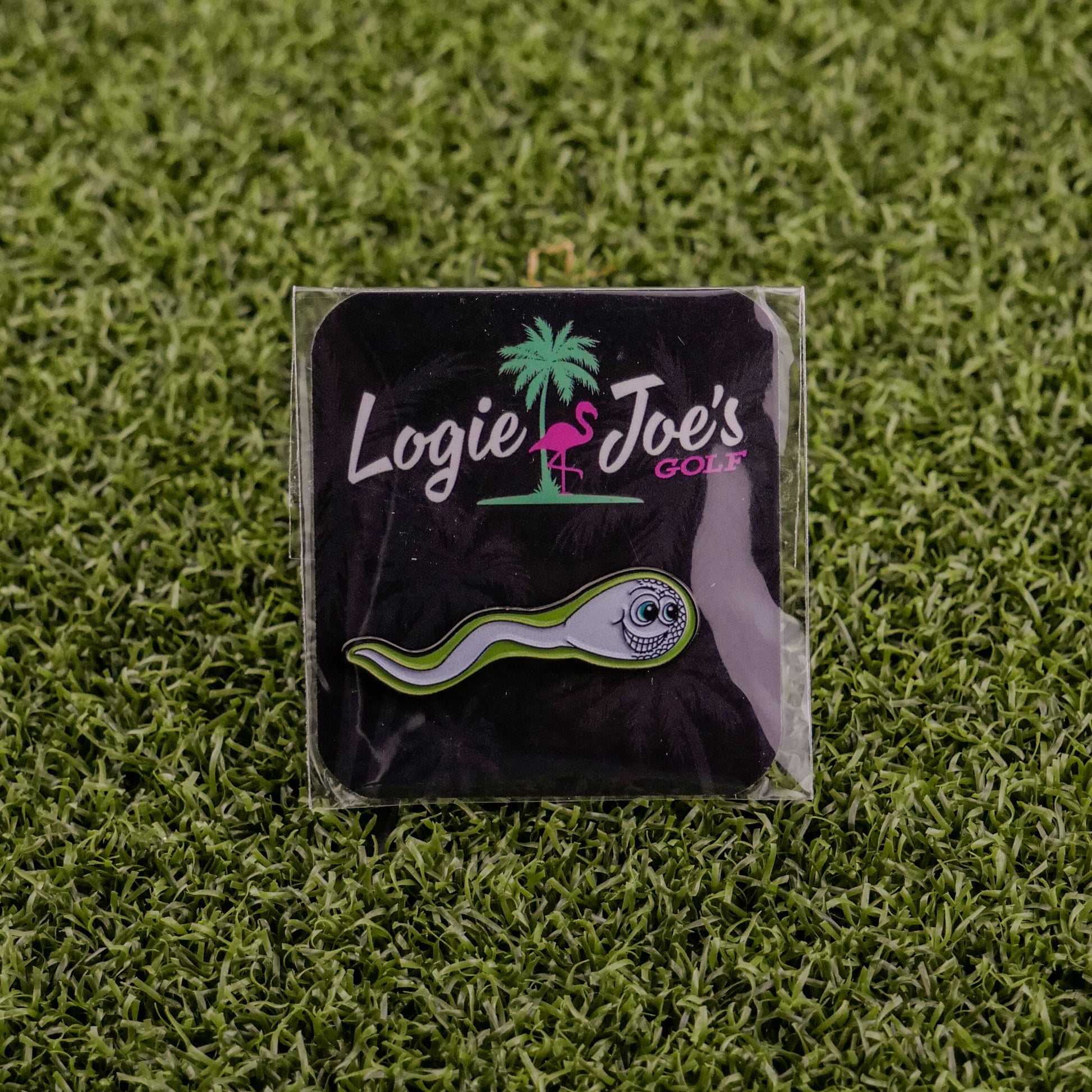 The Sperm Cell, a premium ball marker from Ballmarkers by Logie Joes, features cartoon eyes and comes on artificial grass with a black card in white, green, and pink plus flamingo and palm tree—funny golf accessories at their best.