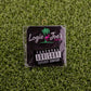 The Parental Advisory sticker by Ballmarkers from Logie Joes features a pink flamingo logo and sits on artificial grass, surrounded by golf ball markers and divot tools.