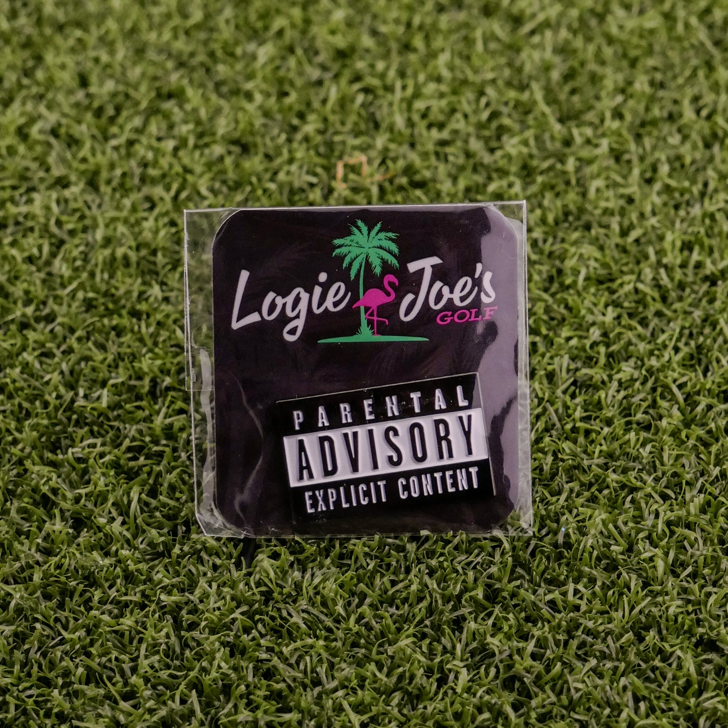 The Parental Advisory sticker by Ballmarkers from Logie Joes features a pink flamingo logo and sits on artificial grass, surrounded by golf ball markers and divot tools.