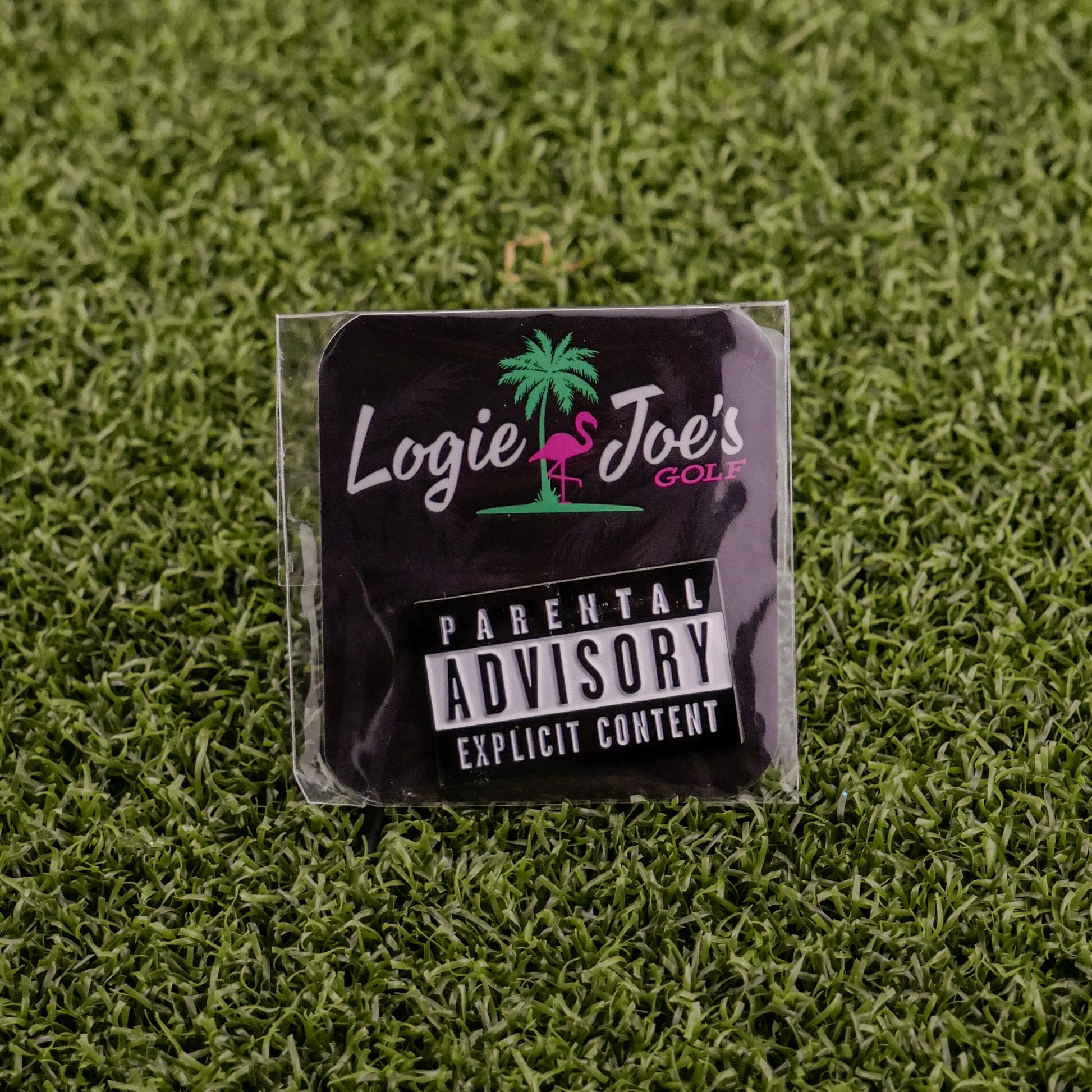 The Parental Advisory sticker by Ballmarkers from Logie Joes features a pink flamingo logo and sits on artificial grass, surrounded by golf ball markers and divot tools.