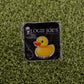 The Rubber Duck You pin by Ballmarkers from Logie Joes features a yellow duck and “DUCK YOU!” text on black packaging, displayed on green turf—ideal for those seeking funny or unique premium golf accessories.