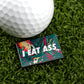 A golf ball on artificial grass sits beside Ballmarkers by Logie Joes I Eat *ss Tropical, a rectangular marker with a bright tropical pattern and bold white letters—just one of their many funny golf accessories.