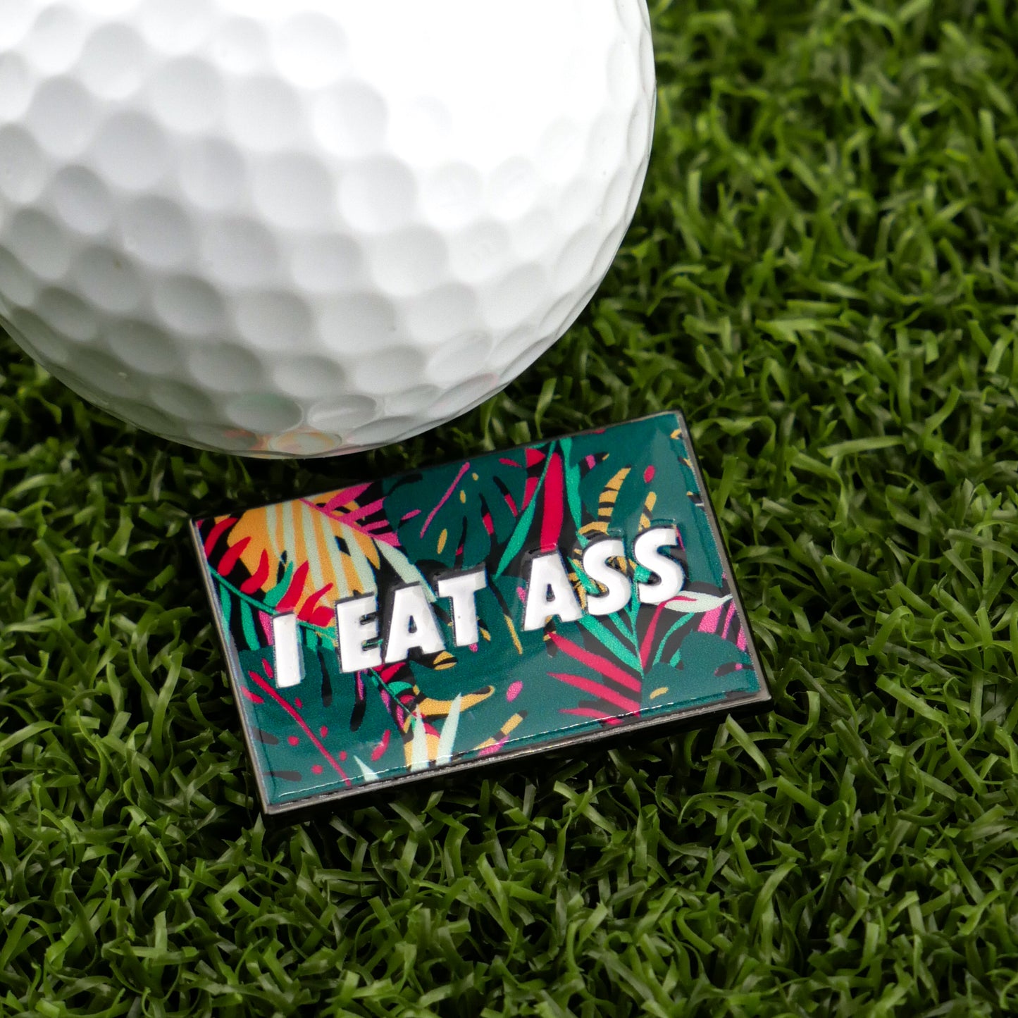A golf ball on artificial grass sits beside Ballmarkers by Logie Joes I Eat *ss Tropical, a rectangular marker with a bright tropical pattern and bold white letters—just one of their many funny golf accessories.