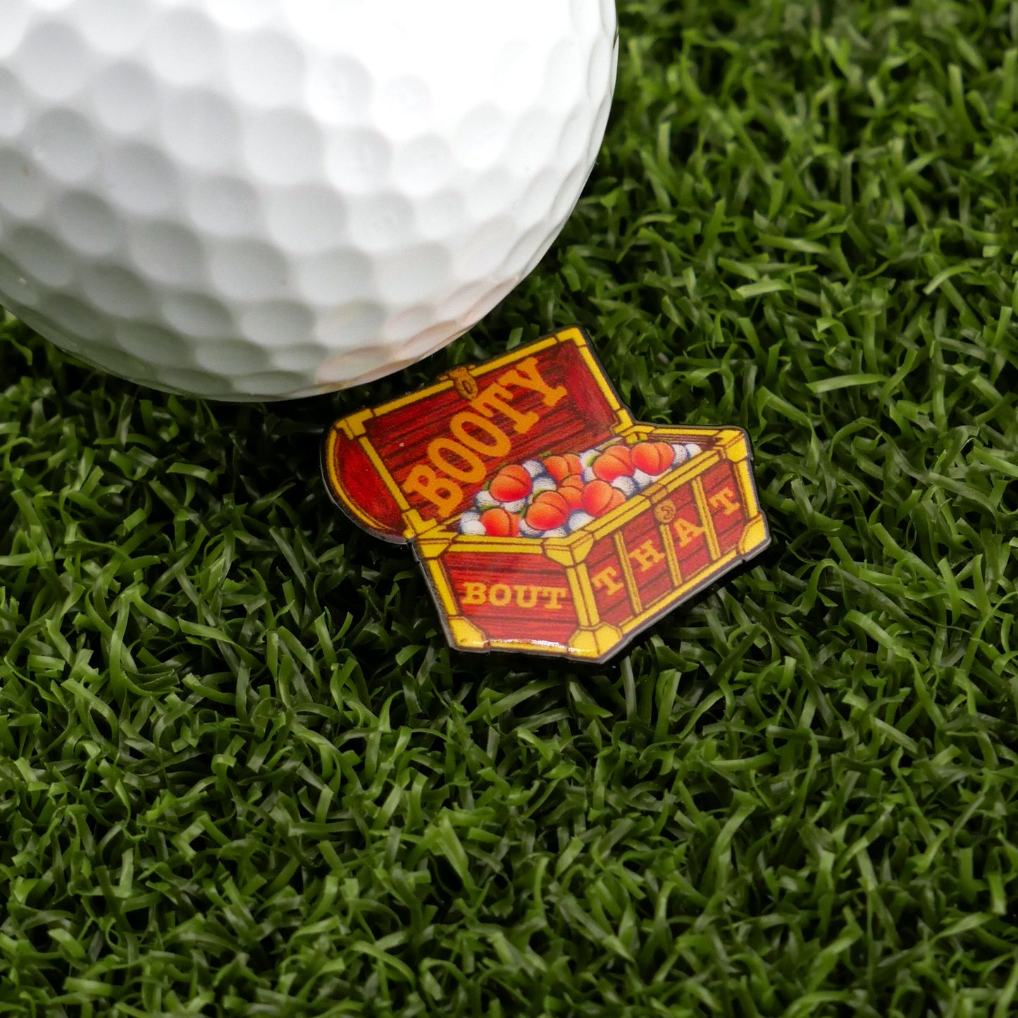A golf ball on artificial grass sits beside the Bout That Booty ball marker from Ballmarkers by Logie Joe’s—a colorful treasure chest with red gems—perfect for fans of unique golf accessories.