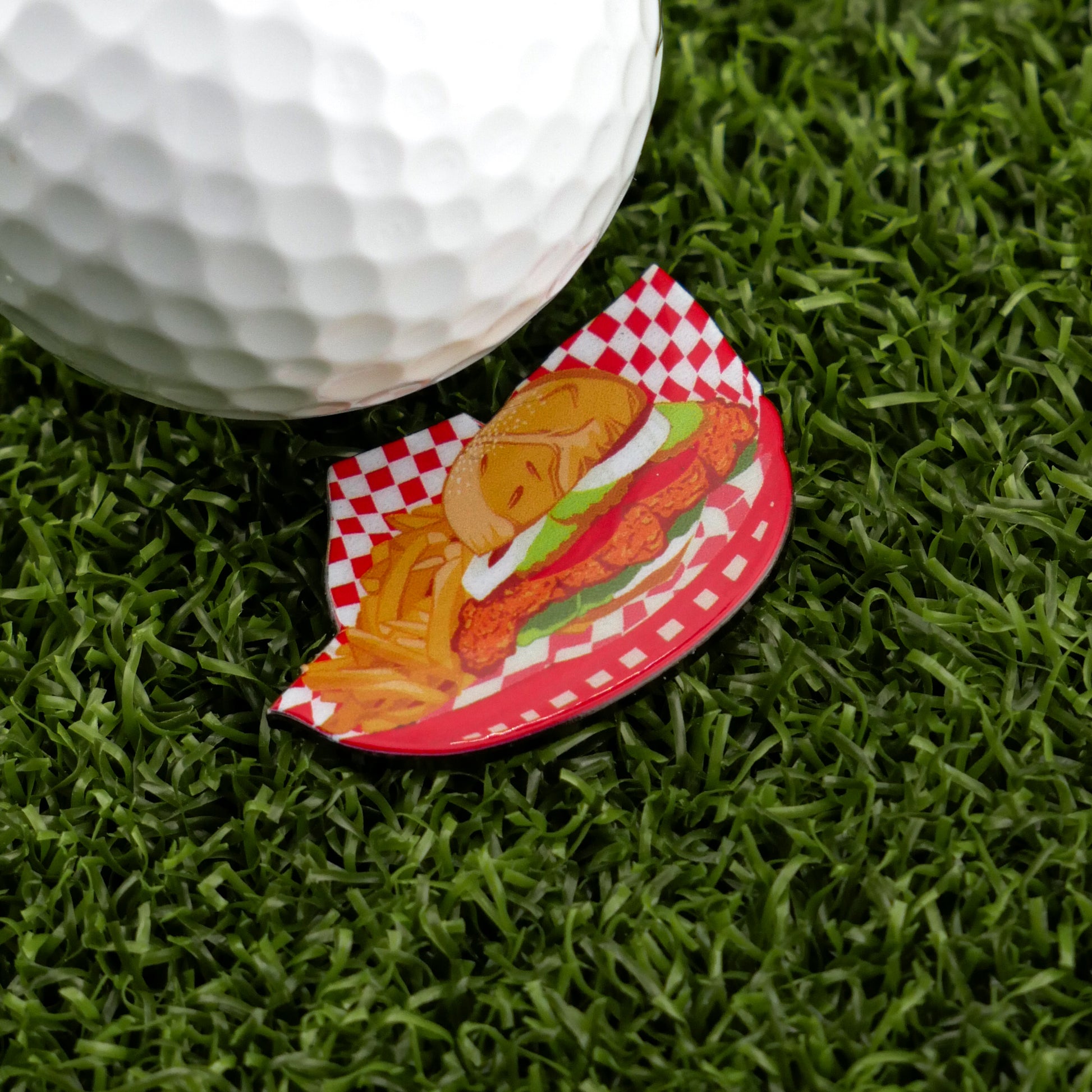 A close-up of a golf ball on artificial grass beside Ballmarkers by Logie Joes Breaded Tenderloin Basket—a premium, humorous ball marker featuring a fried chicken sandwich and fries on a red checkered tray.
