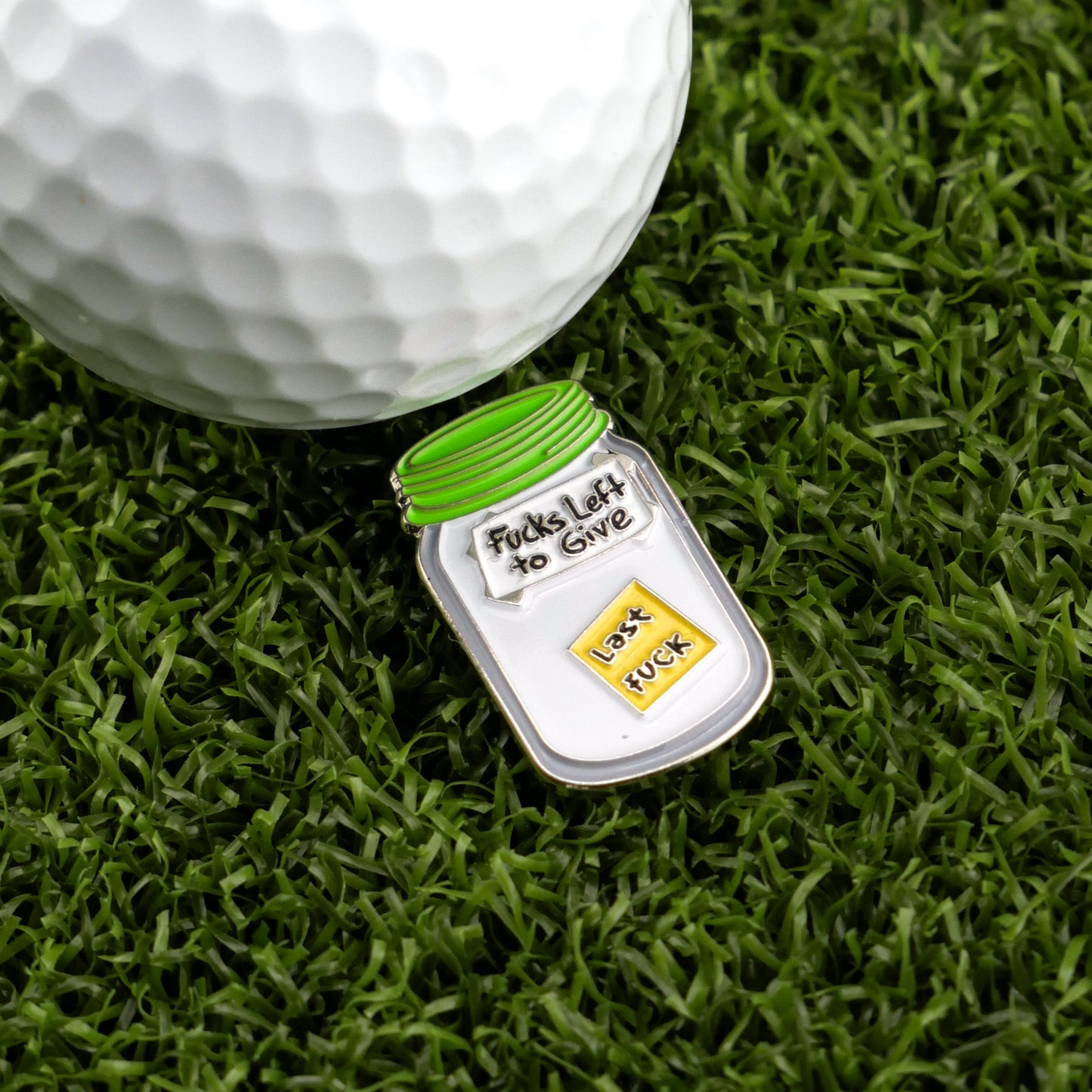 A white golf ball rests on grass beside Ballmarkers by Logie Joe’s Jar of “F”s, a funny ball marker shaped like a jar with a green lid and a single yellow block labeled “last fuck” inside.