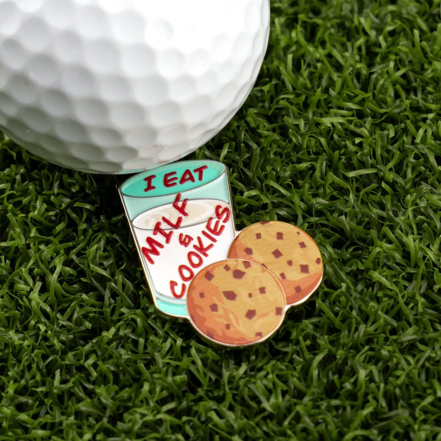 A golf ball on grass beside Ballmarkers by Logie Joe’s “Milf & Cookies” premium marker, featuring a glass of milk labeled “I eat milk & cookies,” with two chocolate chip cookies at its base.