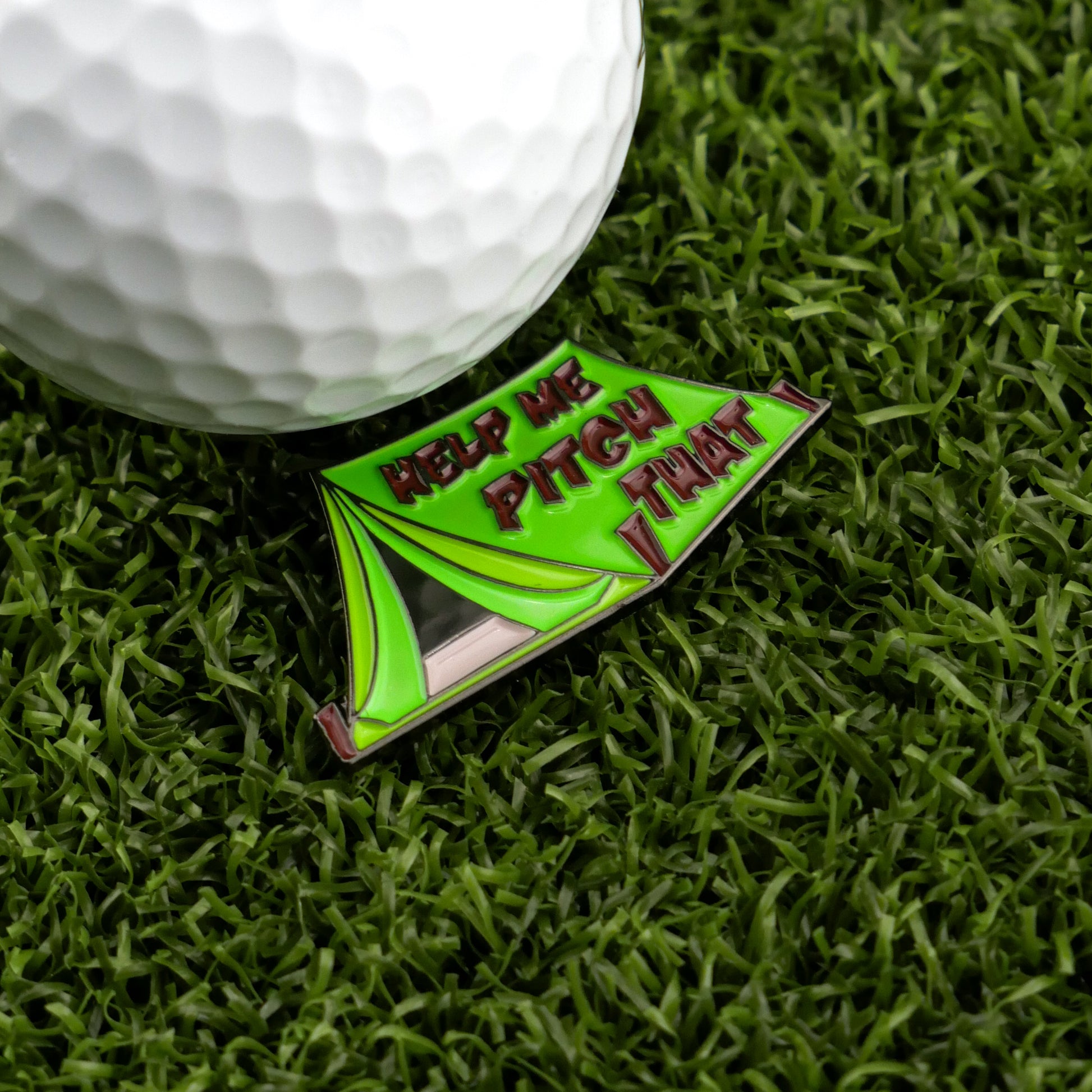 A white golf ball sits on artificial grass beside Ballmarkers by Logie Joes Pitch That Tent enamel pin—a funny, premium ball marker shaped like a green tent and perfect for golfers seeking unique accessories.