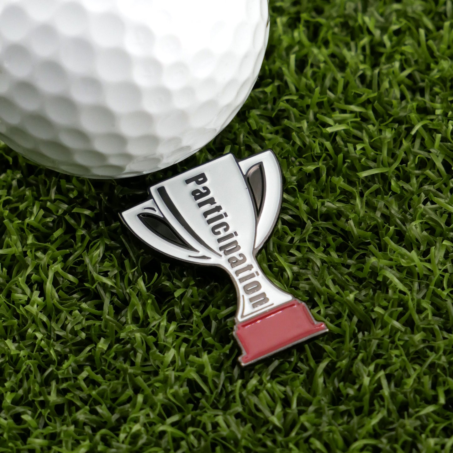 The Participation Trophy by Ballmarkers from Logie Joes, a metal trophy-shaped humorous ball marker, sits next to a white golf ball on artificial grass, bringing a fun twist to premium golf accessories.