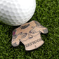 The Titties & Tattoos ball marker from Ballmarkers by Logie Joe’s, shaped like a tattooed upper torso with hands over the chest, sits on green grass by a golf ball—a hilarious gift bound to spark conversation on the course.