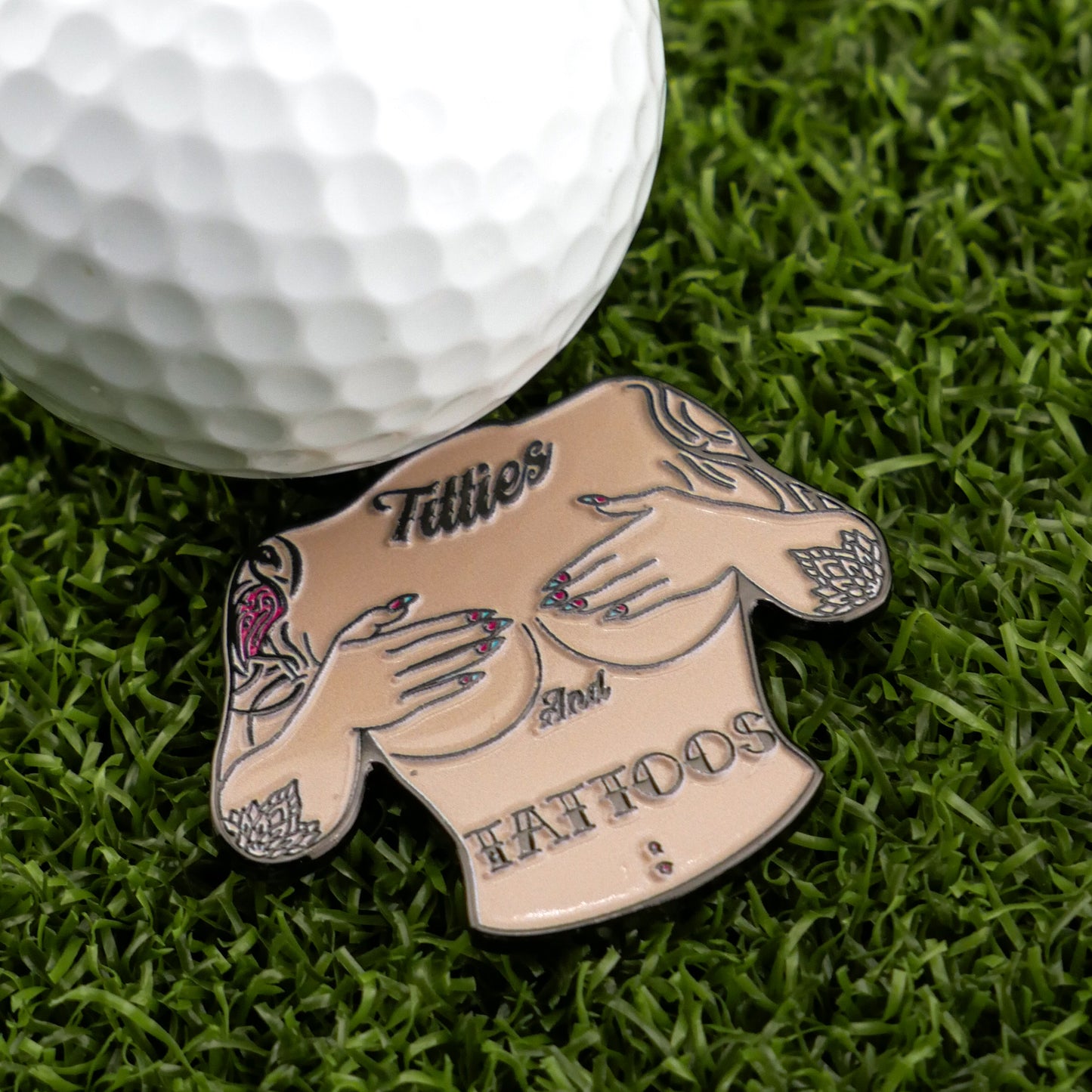 The Titties & Tattoos ball marker from Ballmarkers by Logie Joe’s, shaped like a tattooed upper torso with hands over the chest, sits on green grass by a golf ball—a hilarious gift bound to spark conversation on the course.
