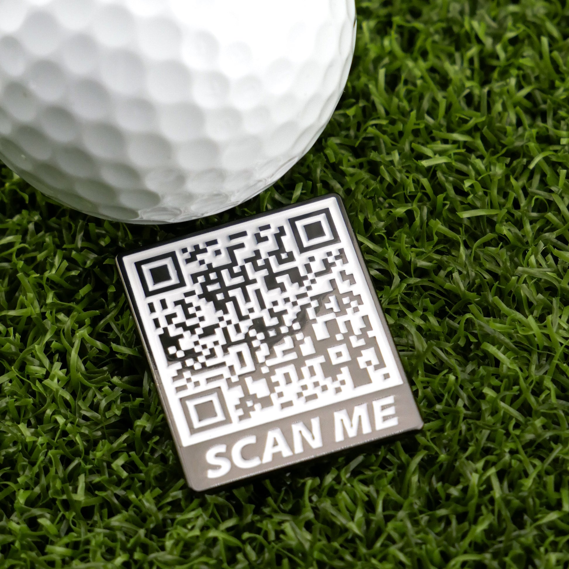 A close-up of a golf ball on artificial grass beside a Ballmarkers by Logie Joe’s Scan Me QR code badge labeled SCAN ME at the bottom.