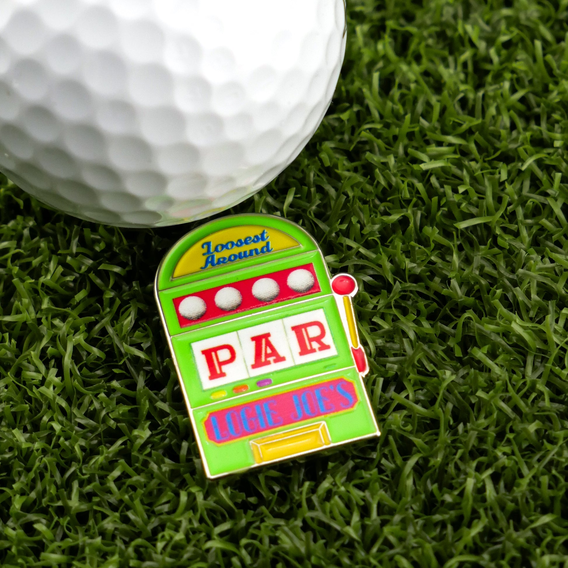 A golf ball on artificial grass sits beside Ballmarkers by Logie Joes Slot Machine, a colorful, slot machine-themed golf ball marker labeled Loosest Around, PAR, and LOGIE JOES—one of the most unique and humorous golf accessories available.