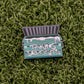 Ball markers by Logie Joes: Green enamel pin shaped like a dumpster with open lid, “TAKING DEPOSITS” on the front, displayed on green artificial grass.