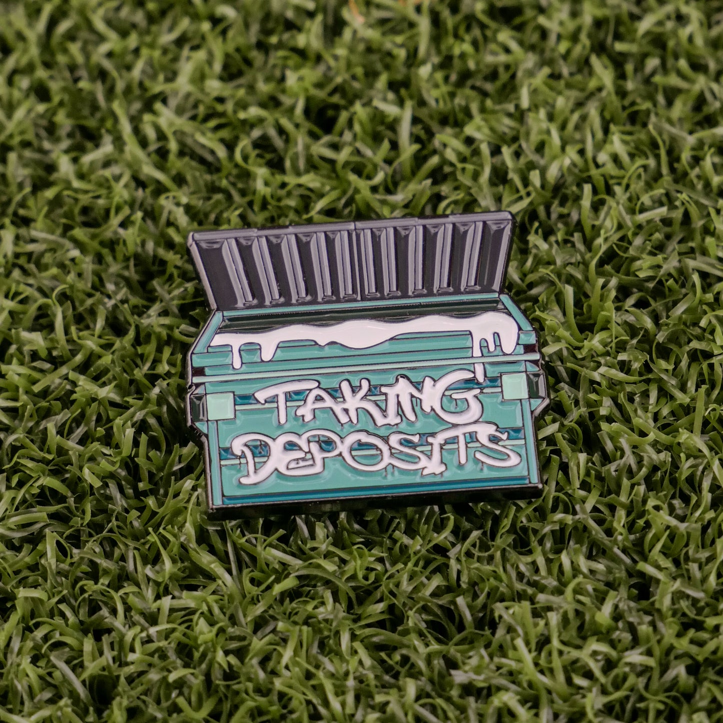 Ball markers by Logie Joes: Green enamel pin shaped like a dumpster with open lid, “TAKING DEPOSITS” on the front, displayed on green artificial grass.
