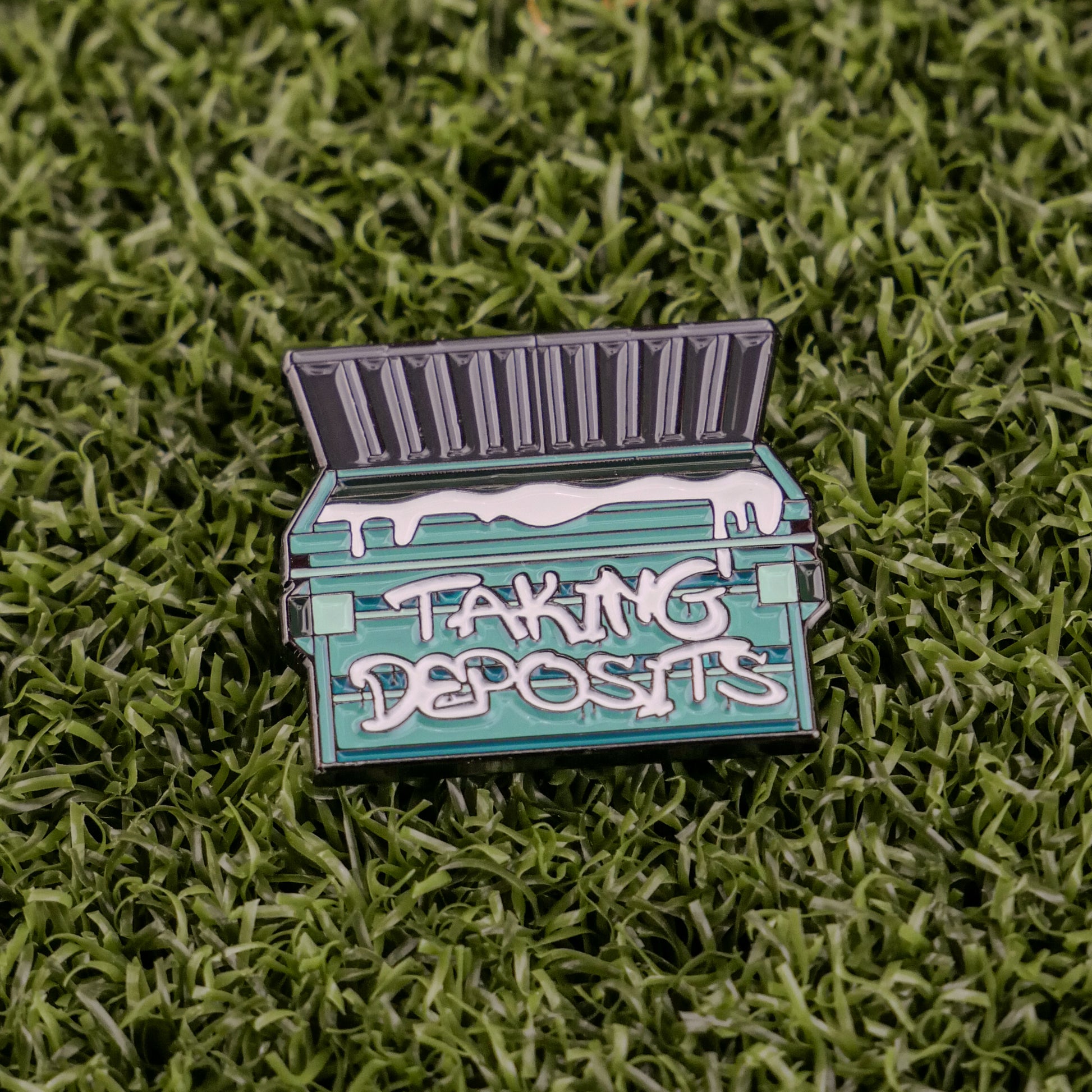 Ball markers by Logie Joes: Green enamel pin shaped like a dumpster with open lid, “TAKING DEPOSITS” on the front, displayed on green artificial grass.