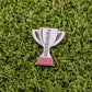 A small silver trophy pin from Ball markers by Logie Joes with Participation inscribed rests on green artificial grass.