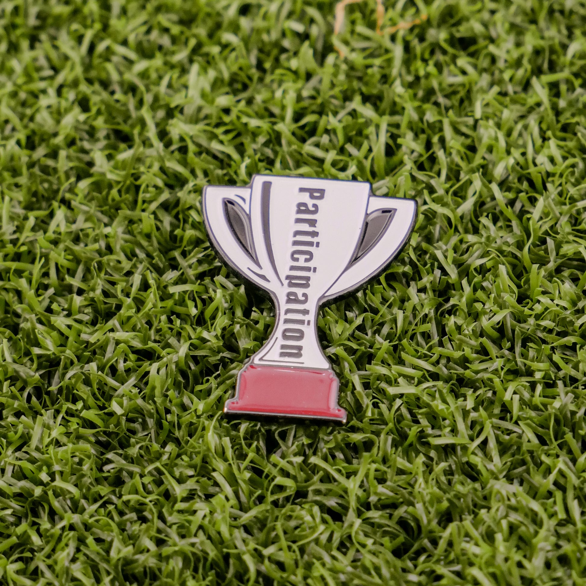 A small silver trophy pin from Ball markers by Logie Joes with Participation inscribed rests on green artificial grass.