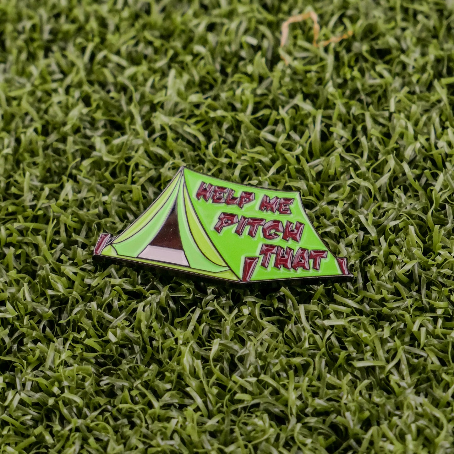 Ball markers by Logie Joes: A green enamel ball marker shaped like a pitched tent with HELP ME PITCH THAT in bold red letters, shown on artificial green grass.