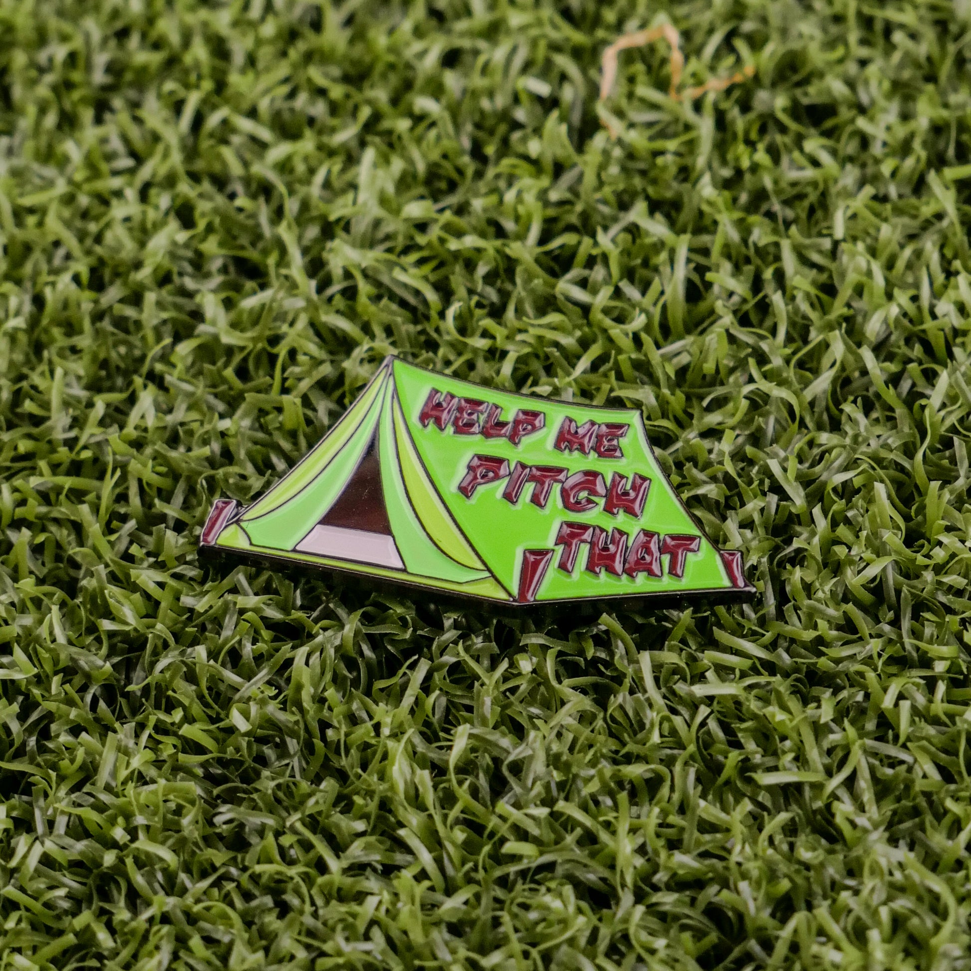 Ball markers by Logie Joes: A green enamel ball marker shaped like a pitched tent with HELP ME PITCH THAT in bold red letters, shown on artificial green grass.