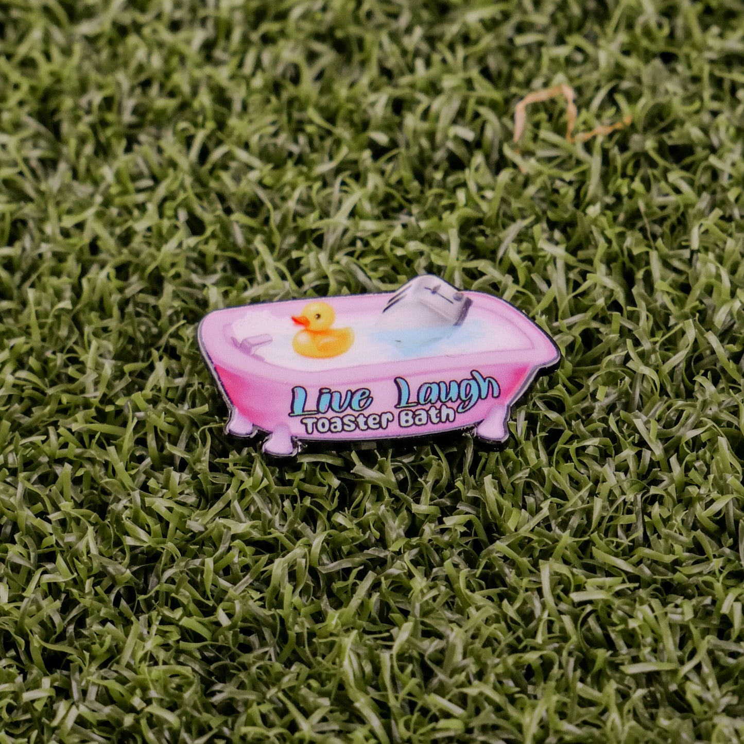 A pink bathtub pin from Ball markers by Logie Joes features a rubber duck, toaster, and the words Live Laugh Toaster Bath, displayed on green artificial grass.
