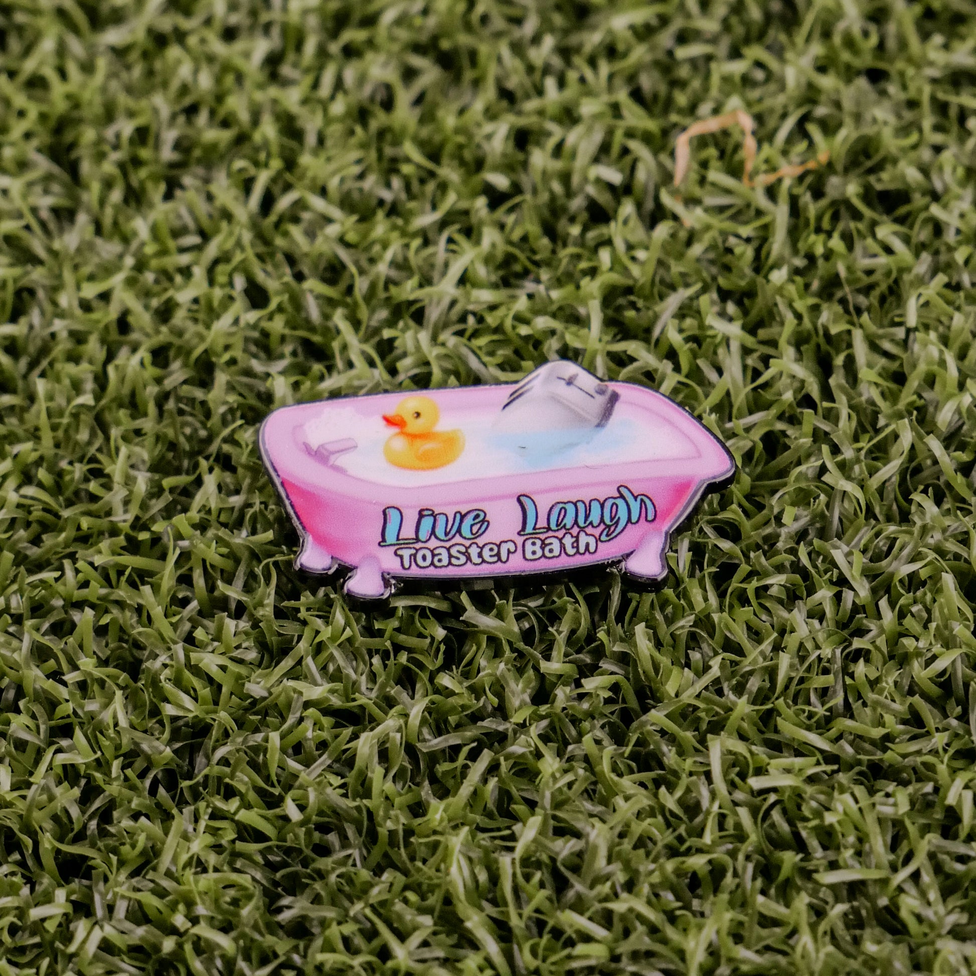 A pink bathtub pin from Ball markers by Logie Joes features a rubber duck, toaster, and the words Live Laugh Toaster Bath, displayed on green artificial grass.