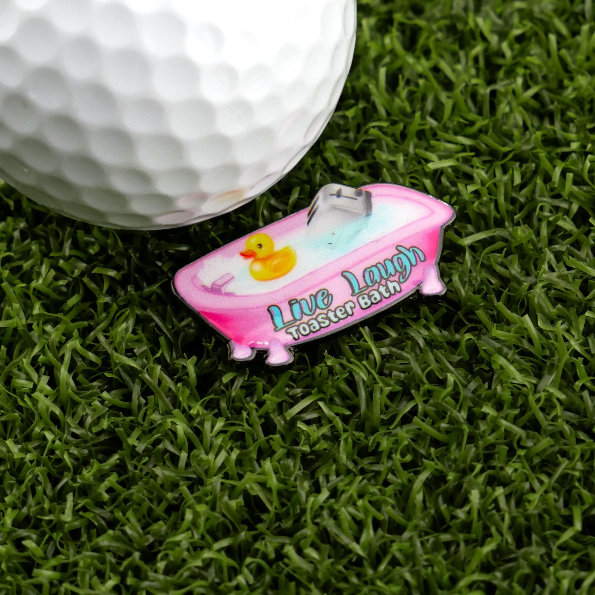 A golf ball on green grass sits beside Ballmarkers by Logie Joes Live Laugh Toaster Bath—a pink enamel golf ball marker shaped like a bathtub with a yellow rubber duck and witty lettering.